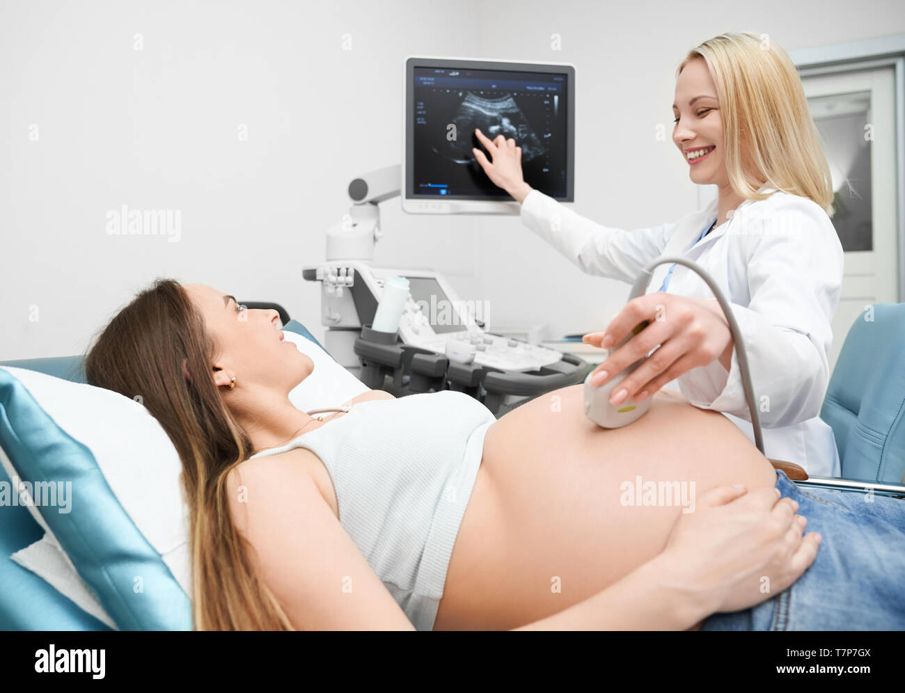 Professional female doctor pointing at screen and showing little baby to future mother. Pregnant woman lying on couch during ultrasound procedure in medical center. Concept of diagnostic and health. Stock Photo