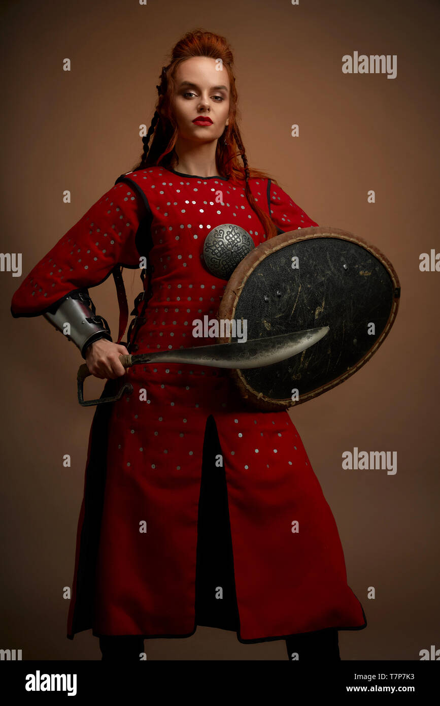 Front view of female gladiator warrior in armor keeping big knife and shield and looking at camera on isolated background in studio. Strong fighter in armor posing. Concept of weapon and power. Stock Photo