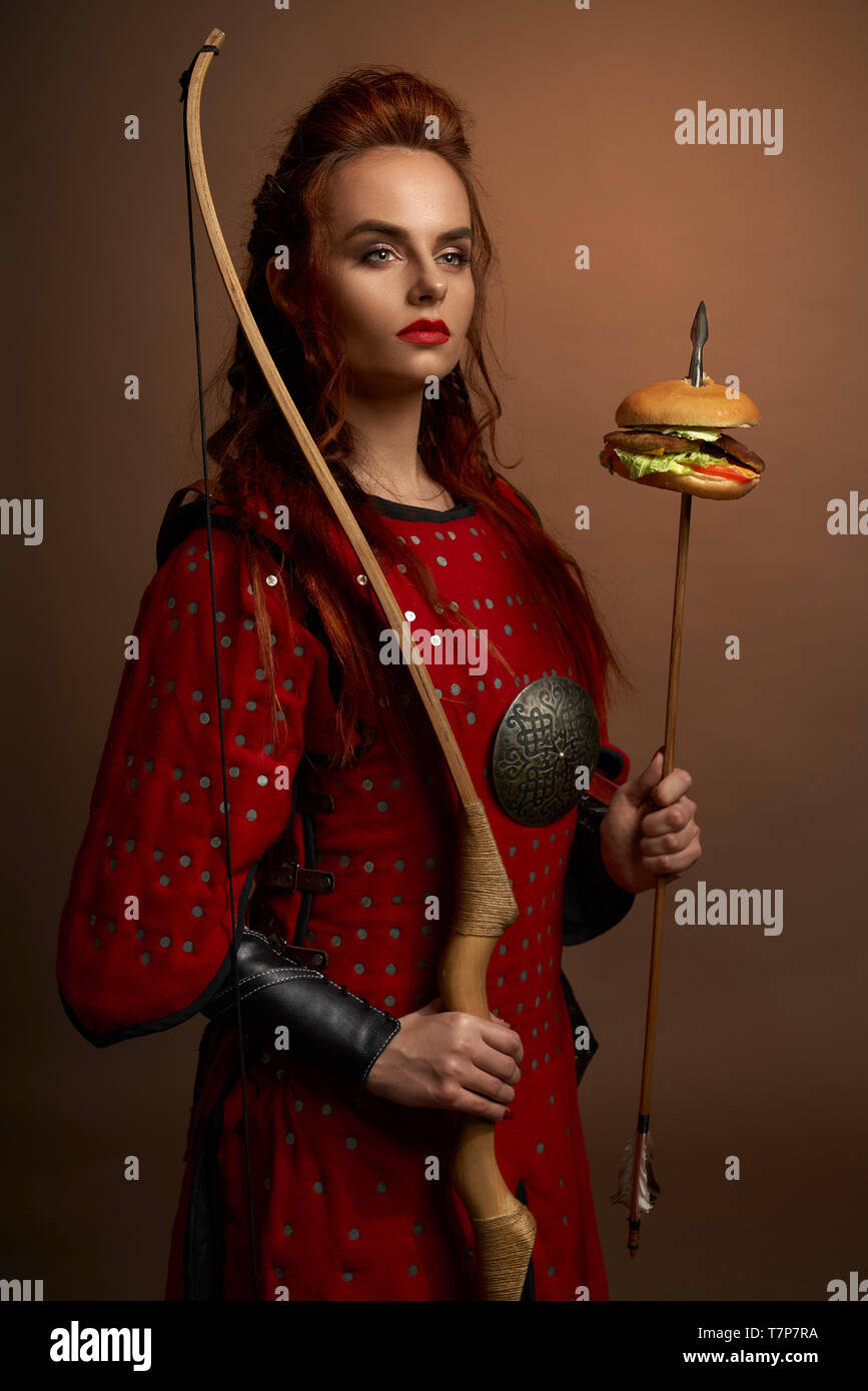 Portrait of determined female fighter in red armor keeping bow and arrow with hamburger in studio. Spartan warrior looking aside and posing on isolated background. Concept of power and food. Stock Photo