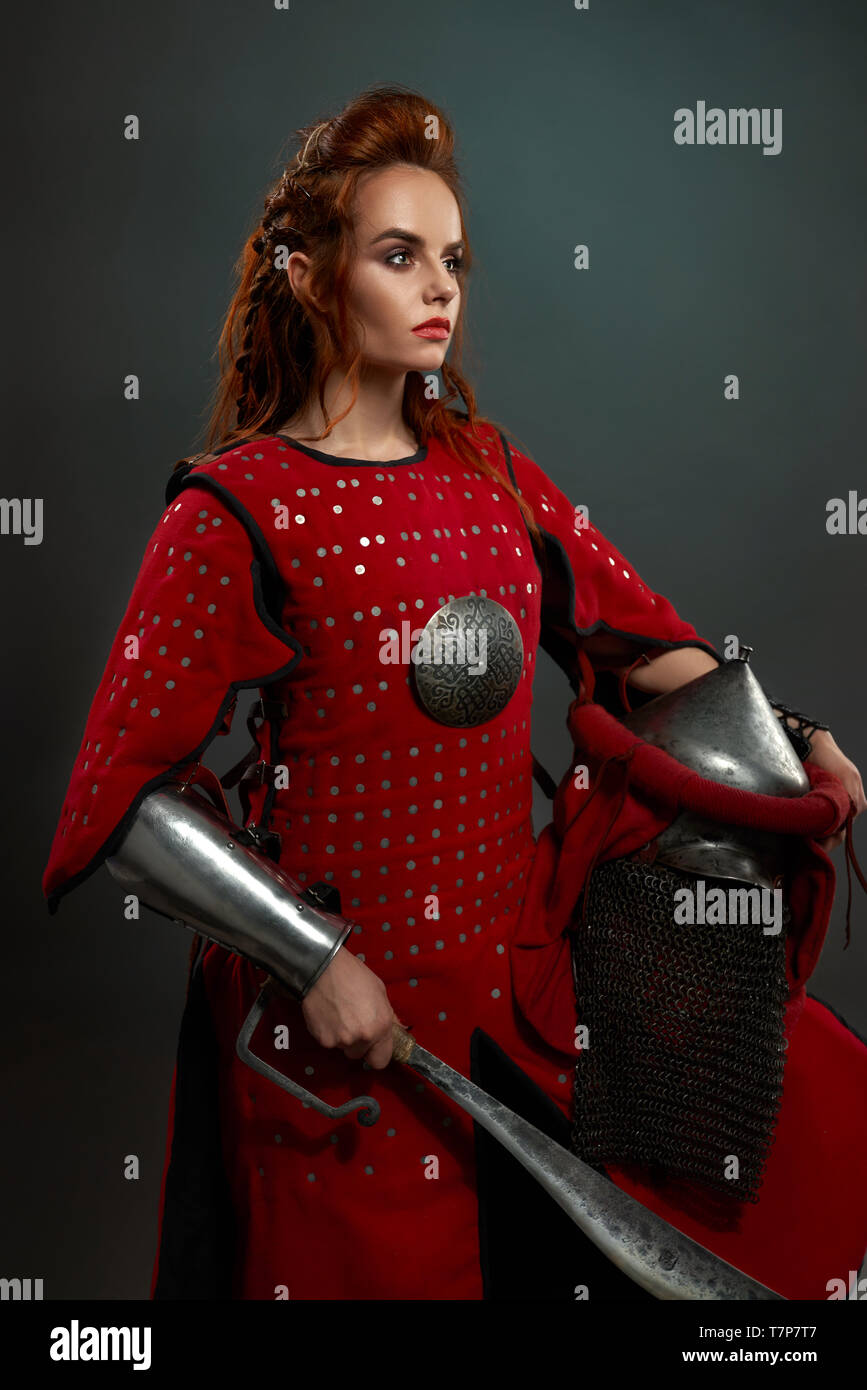 Side view of attractive courageous warrior in red armor keeping helmet and knife and posing on isolated background in studio. Female fighter looking aside and posing. Concept of power and battle. Stock Photo