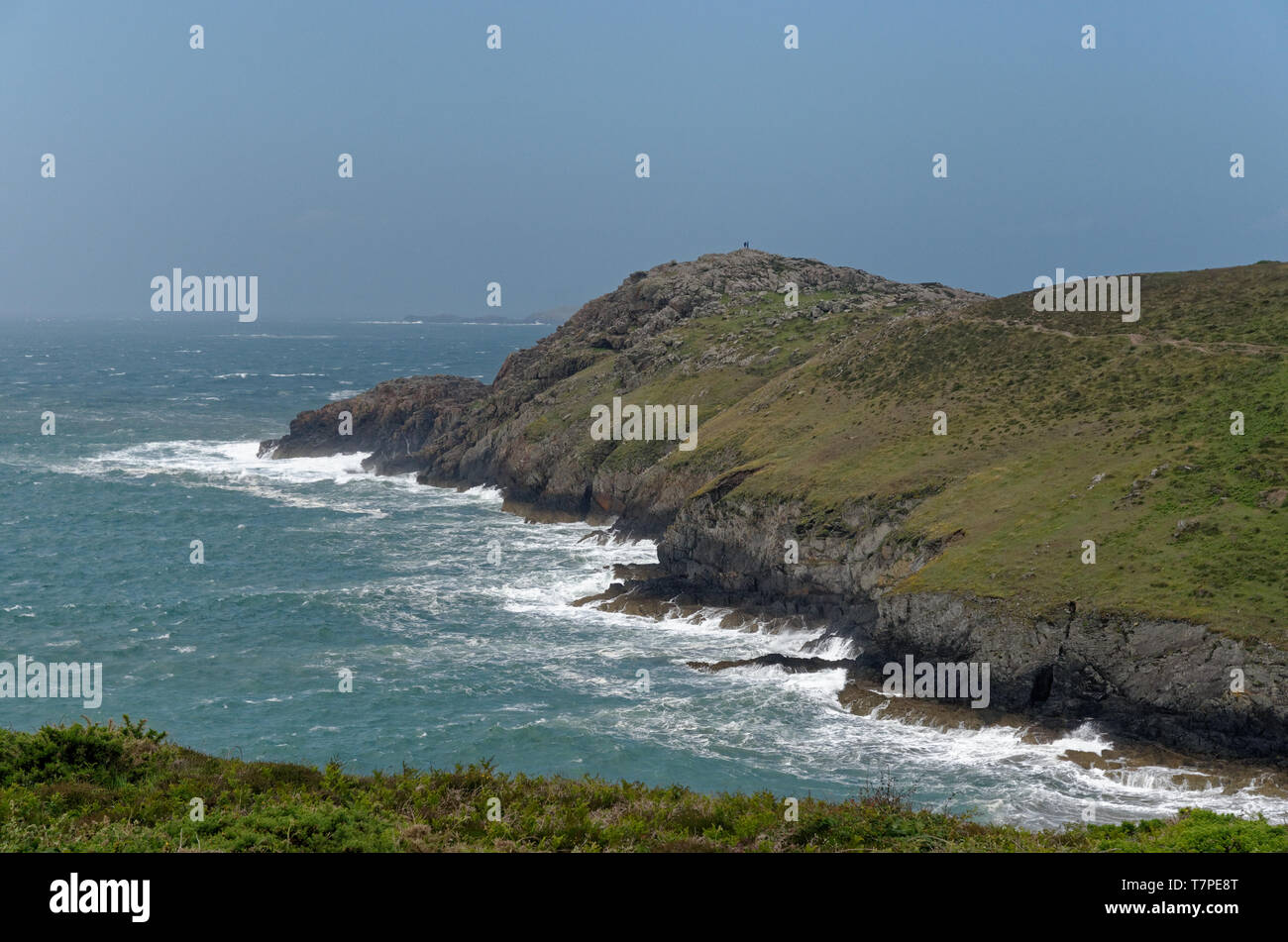 Distant figures on top of a rocky peninsula being pounded by Irish Sea waves on the Pembrokeshire Coast Path near Whitesands Bay in western Wales Stock Photo