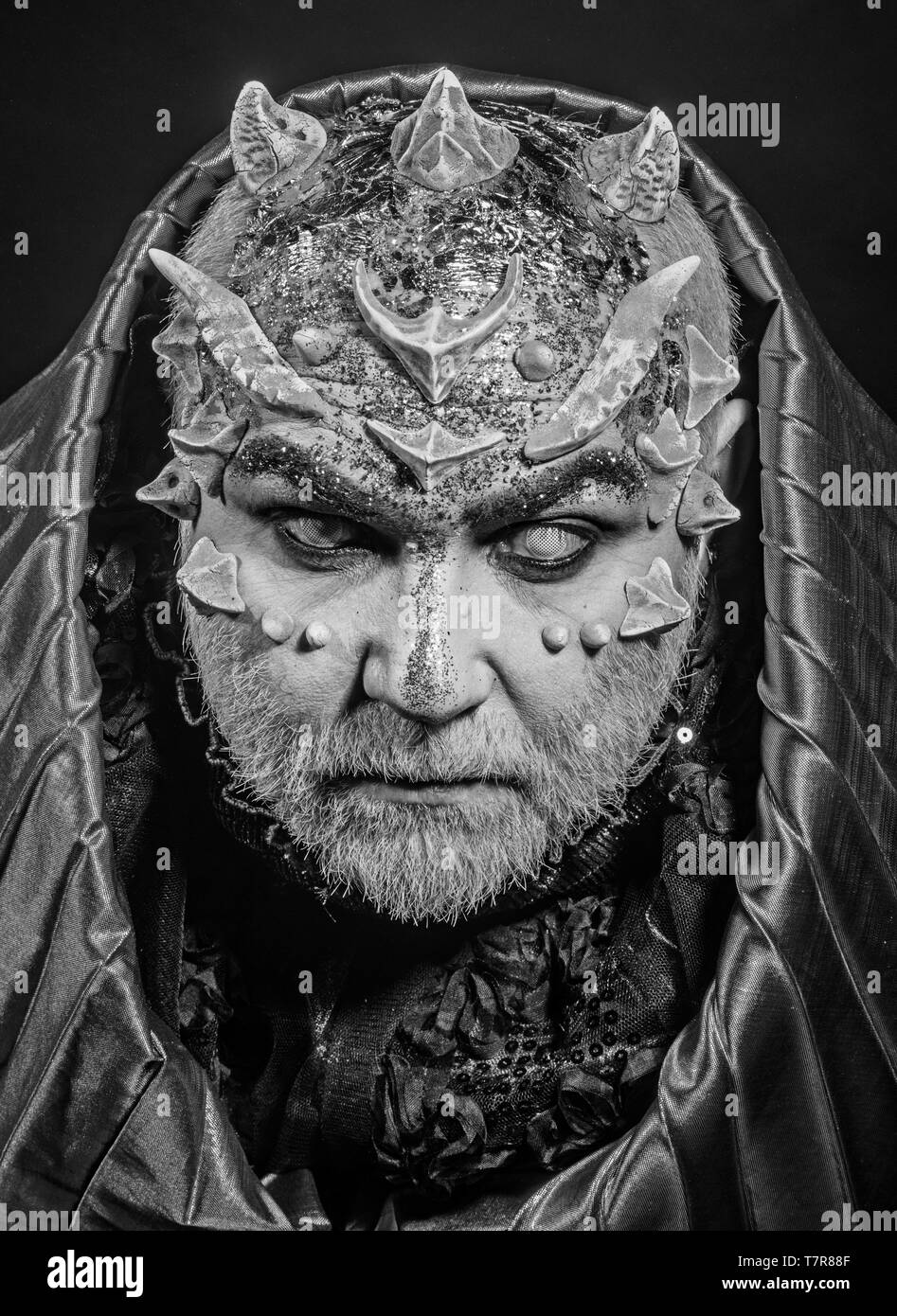 Man with thorns or warts, face covered with glitters. Demon with golden hood on black background. Senior man with white beard dressed like monster Stock Photo