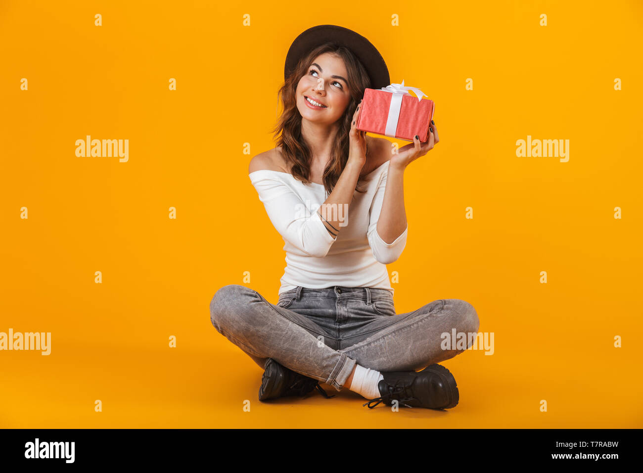 Portrait of a cheerful young woman wearing white shirt and hat sitting isolated over yellow background, holding gift box Stock Photo