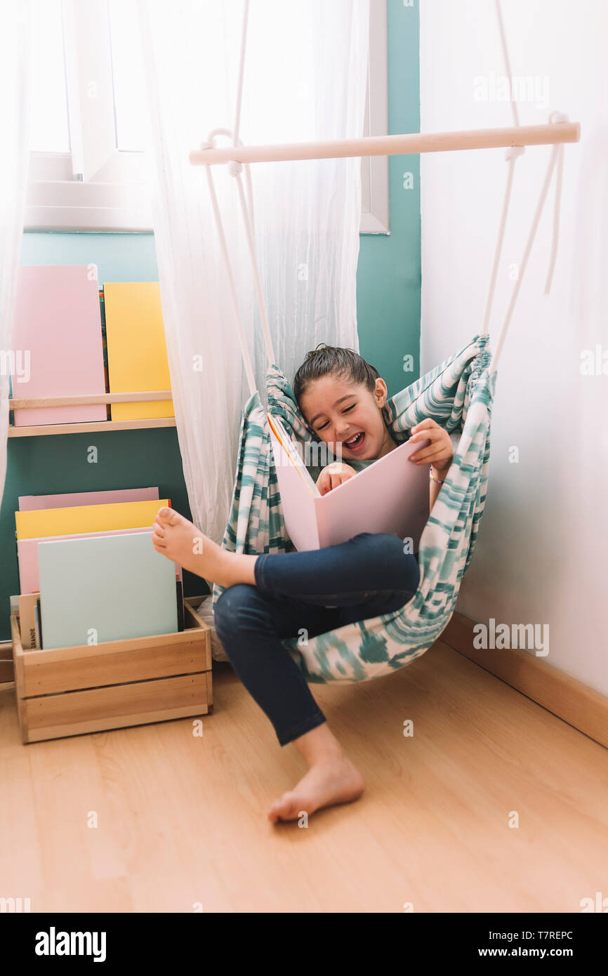 vertical photo of a little girl laughing while reading a book at home ...