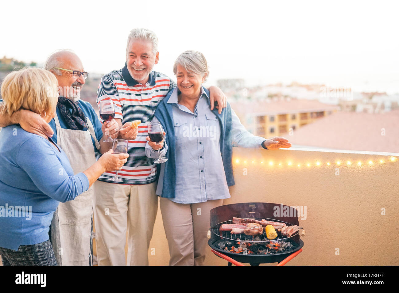 Happy senior friends having fun drinking red wine at barbecue dinner in terrace - Mature people dining and laughing together on rooftop Stock Photo