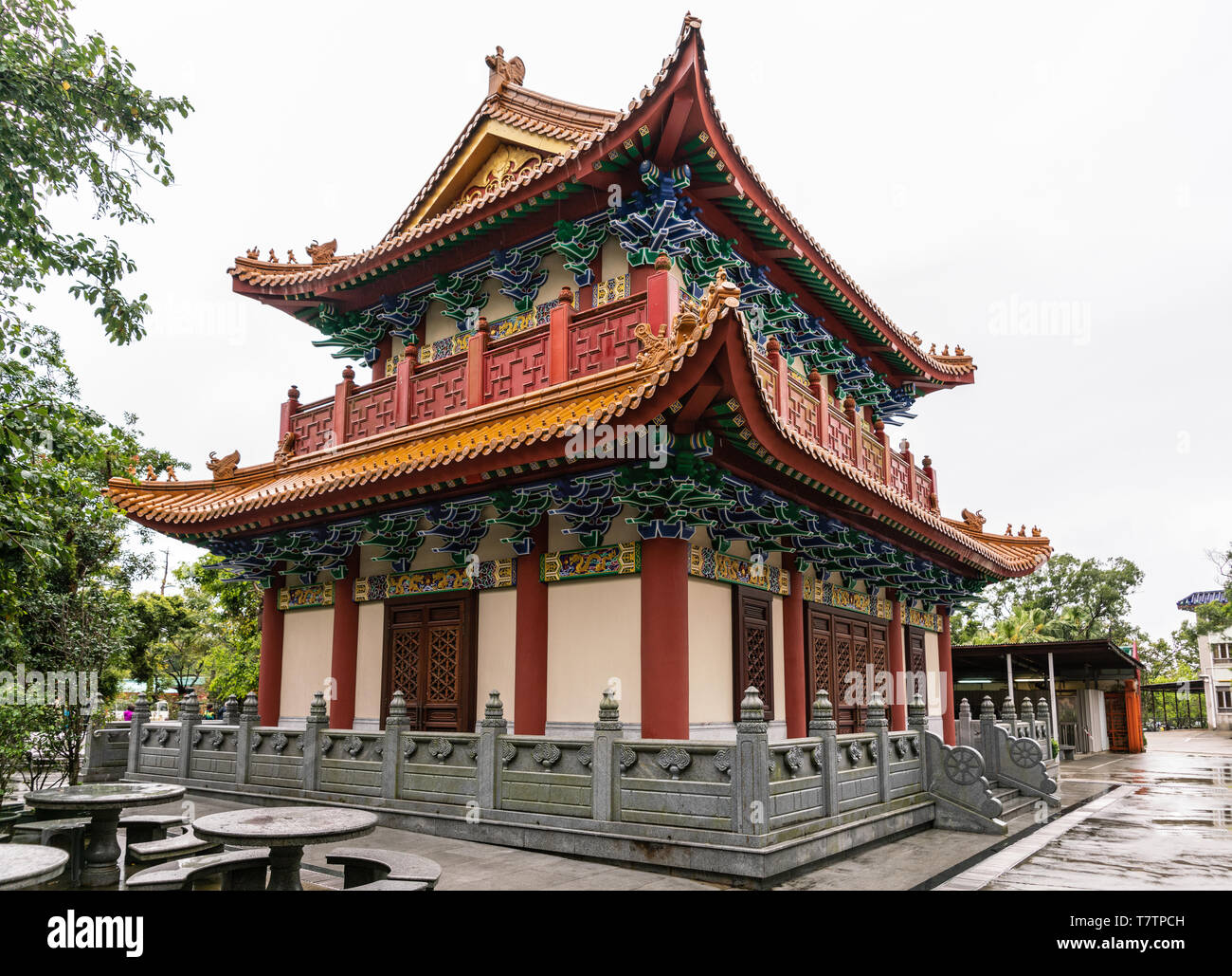 Hong Kong, China - March 7, 2019: Lantau Island. Po Lin Buddhist Monastery. Small red-roofed hall under silver sky. Some green foliage. Decorated wall Stock Photo