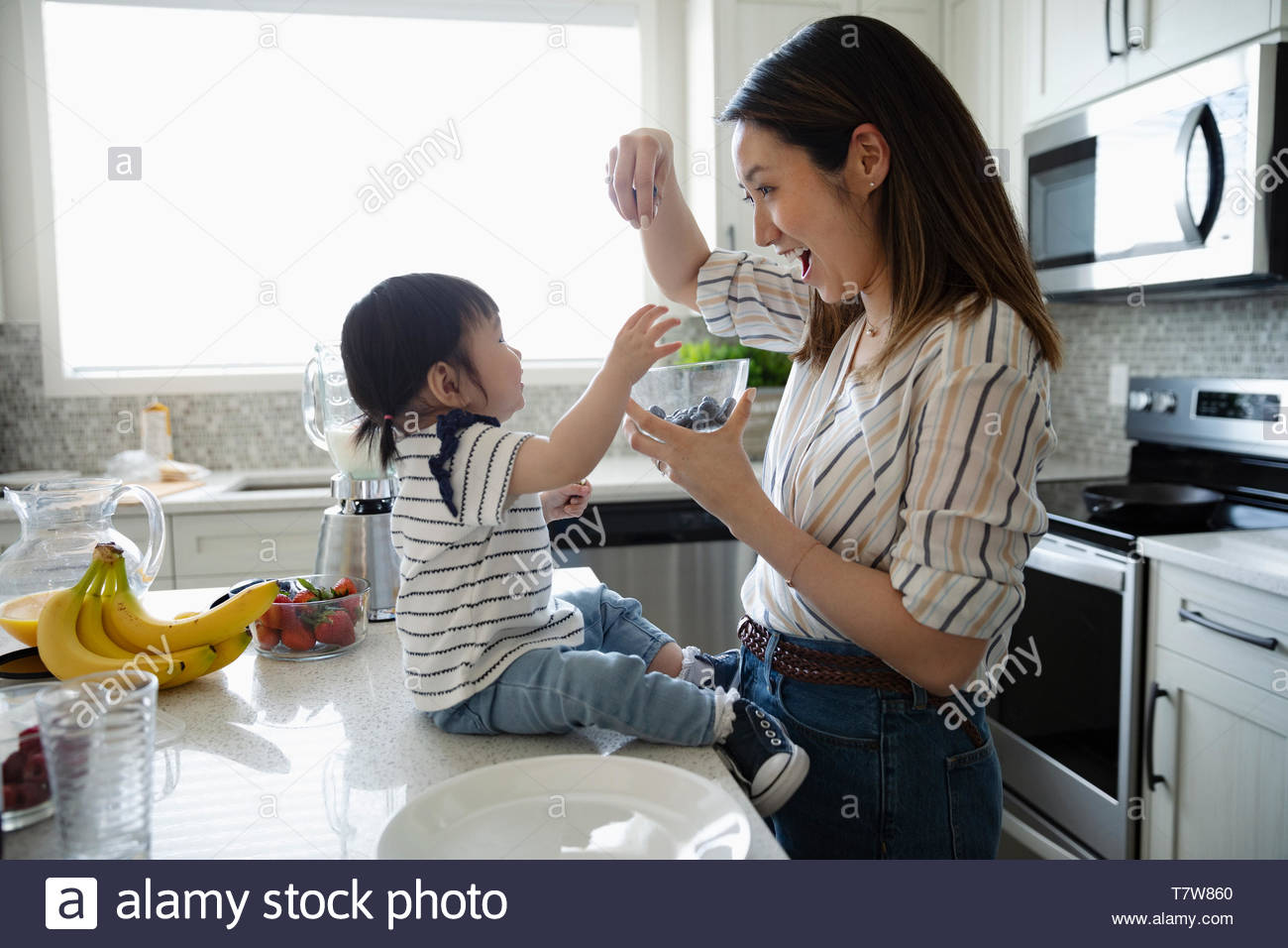 Mother and toddler daughter eating blueberries in kitchen Stock Photo