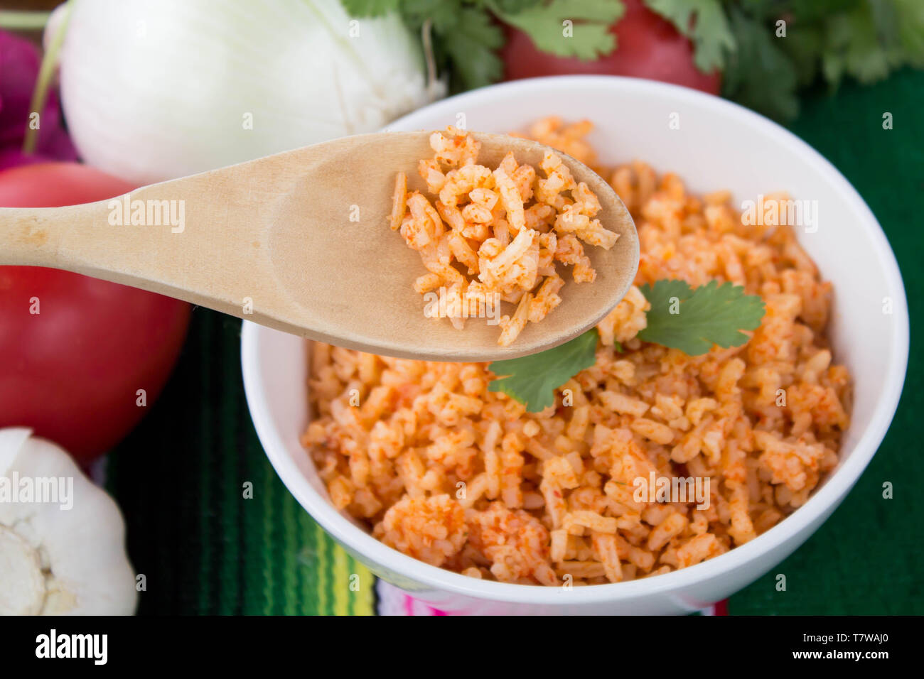 Mexican -style rice in white bowl with close-up of wooden spoonful of rice in foreground, ingredients set against green background Stock Photo