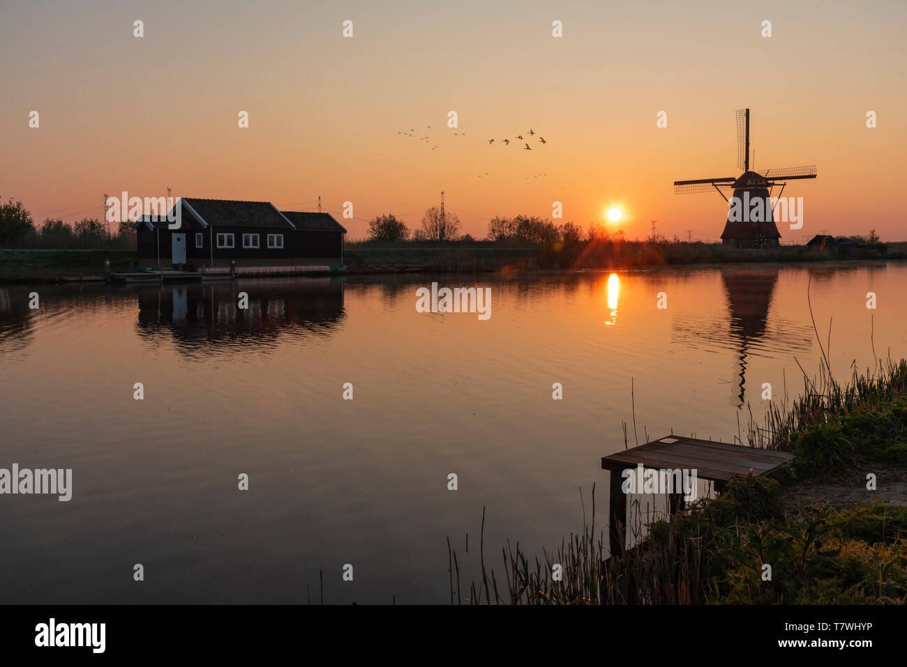 Loading platform at the edge with the calm water in the long canal during facing a windmill reflection in the burning sunrise color morning Stock Photo