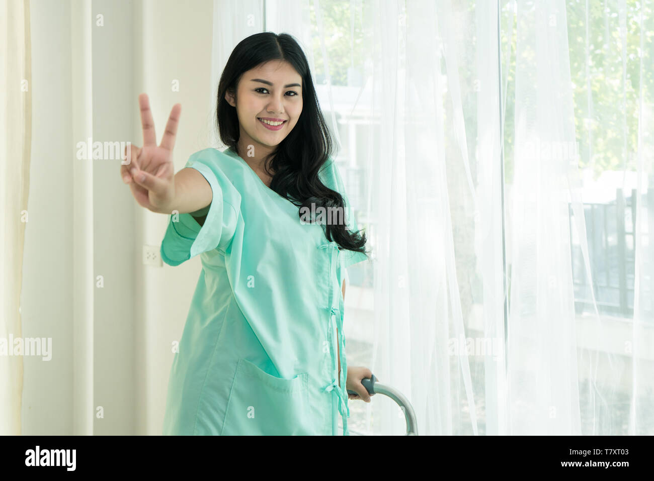 Young Asian patient woman standing at hospital room with walking stick showing victory sign for cheerful. Stock Photo