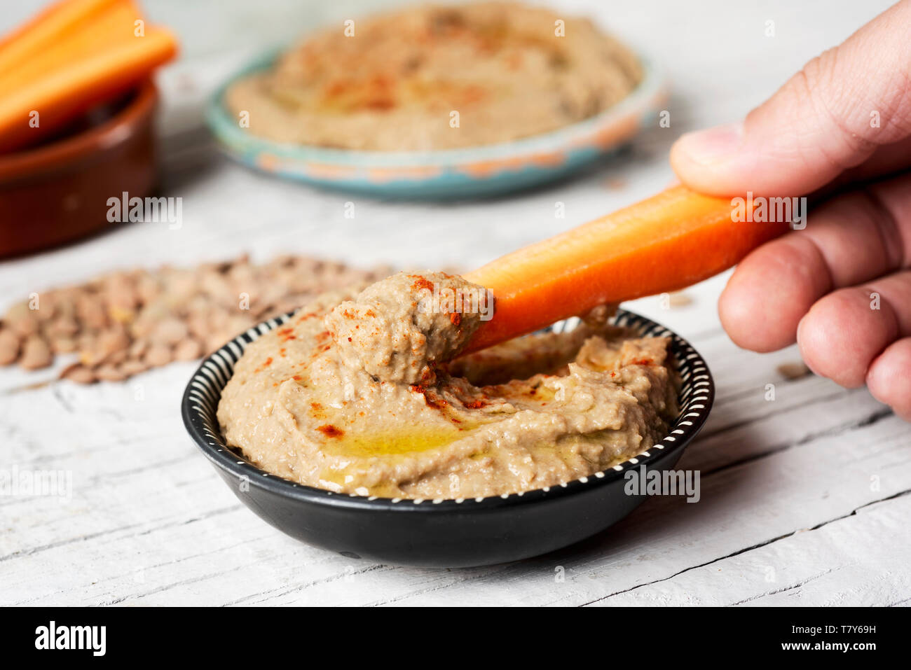 closeup of a man dipping a strip of carrot in a homemade lentil hummus seasoned with paprika served in a black and white plate, on a white rustic wood Stock Photo
