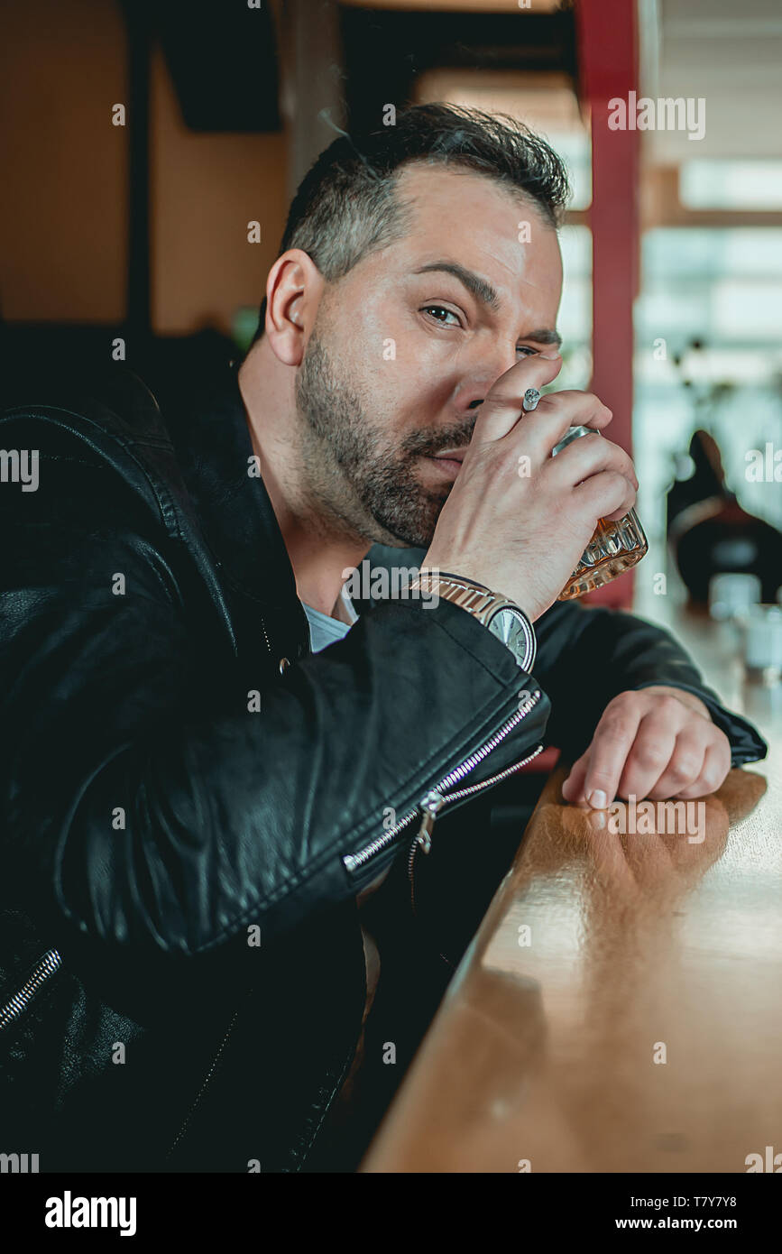 A Man in a bar at the counter looks up at the camera with raised eyebrow and whiskey in his hand Stock Photo