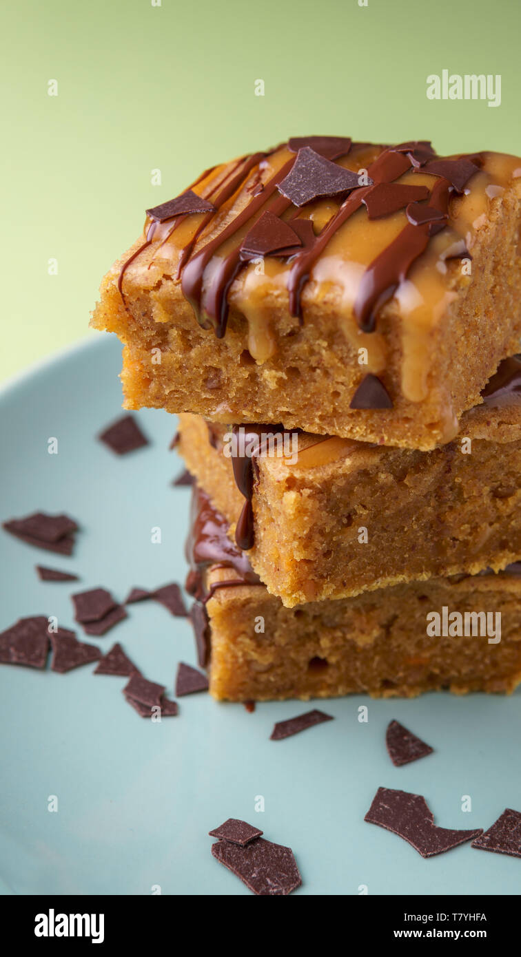 Stack of three sweet potato blondies with thin lines of chocolate and caramel icing running down at the sides and chocolate chips on teal plate, green Stock Photo
