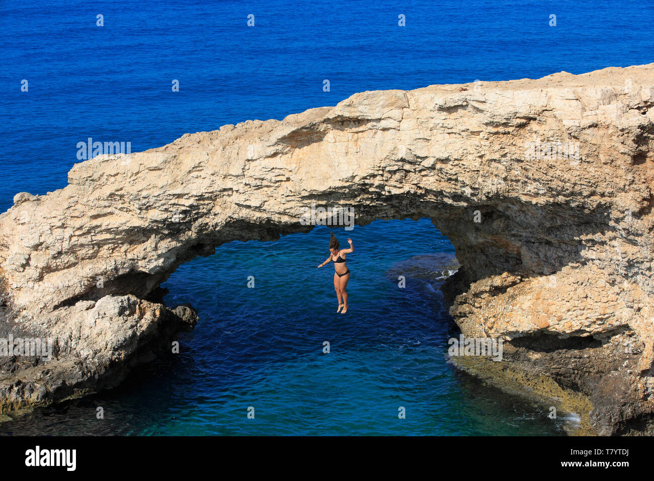 Young girl jumping from the 'Love Bridge', a natural stone arch, into the shallow turquoise water in Ayia Napa, Cyprus Stock Photo