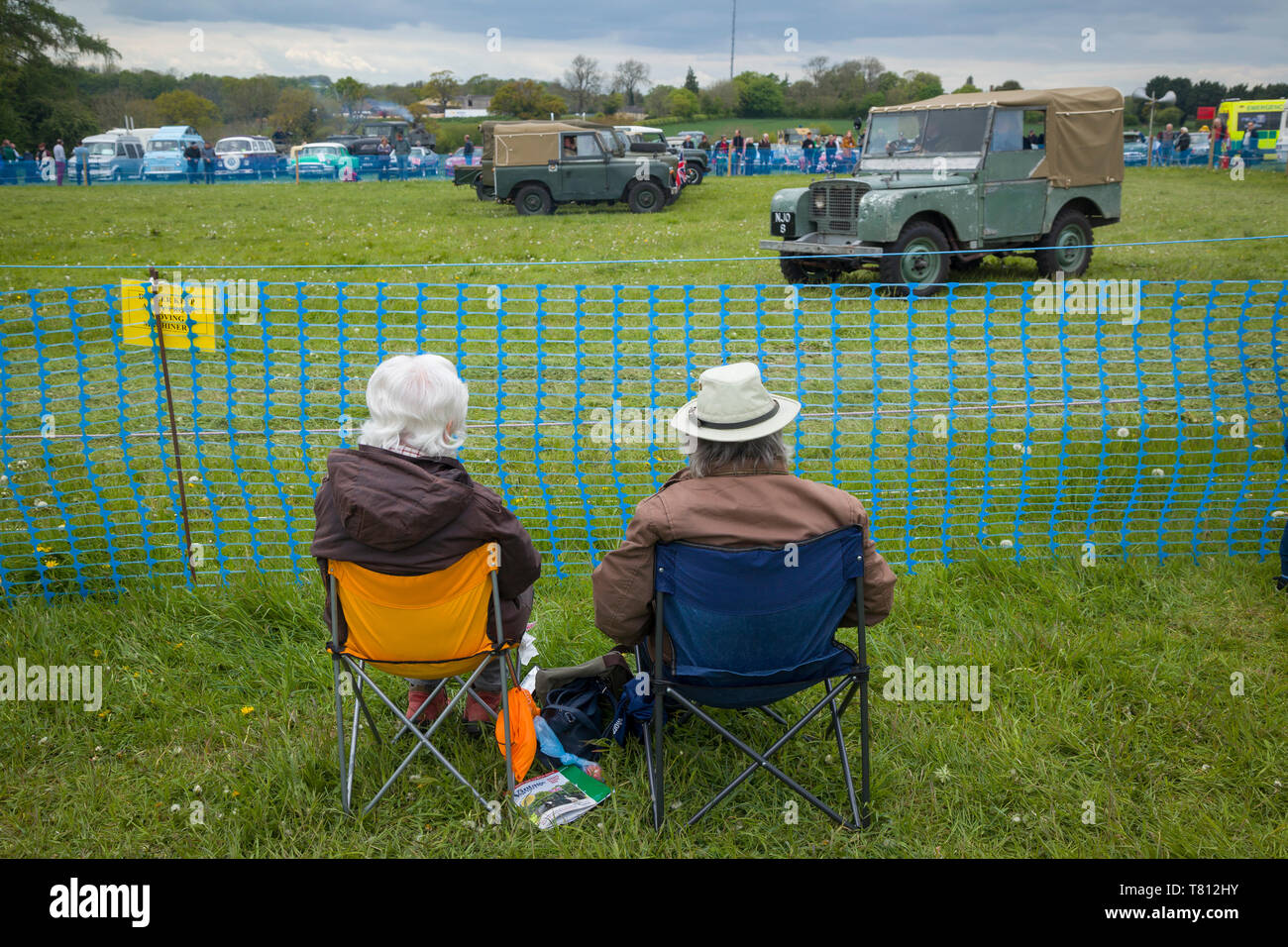 Two seated spectators watch a display of classic Land Rovers at a country show. Stock Photo