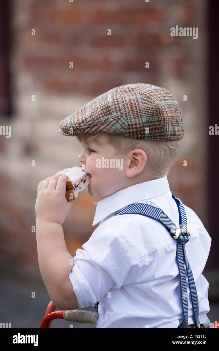 Close up cute little WW2 boy, 1940s costume in flat cap & braces playing outdoors, eating big jam sandwich, 1940 wartime event, UK. Stock Photo