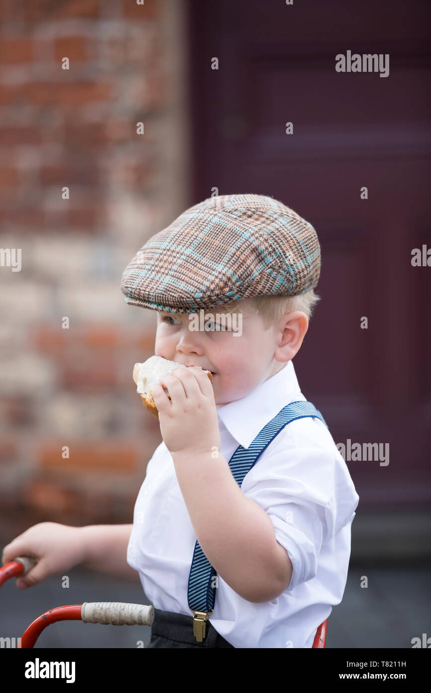 Close up cute little WW2 boy in 1940s costume flat cap & braces isolated outdoors eating big jam sandwich, Black Country Museum, WWII summer event, UK. Stock Photo