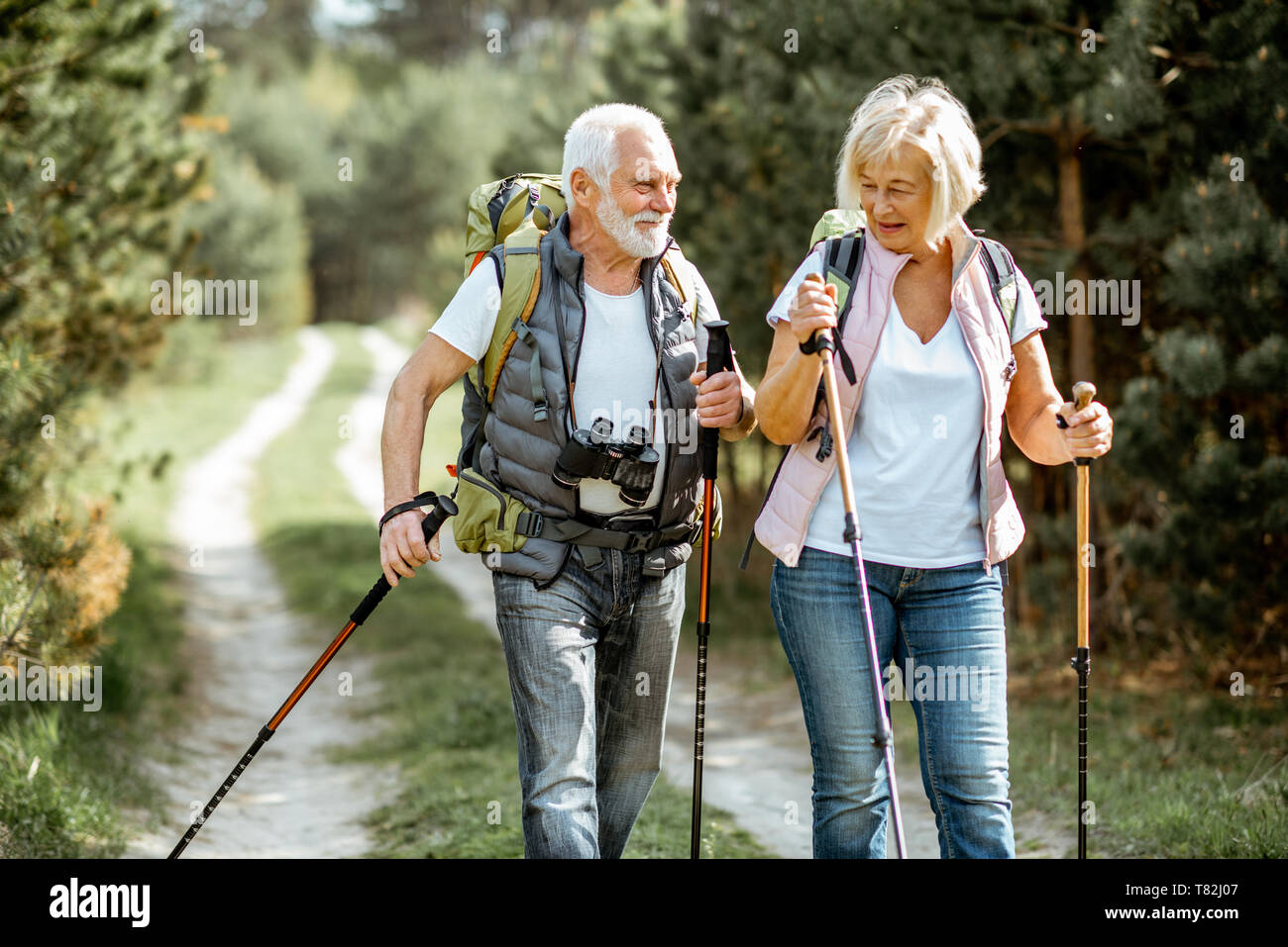 Happy senior couple hiking with trekking sticks and backpacks at the young pine forest. Enjoying nature, having a good time on their retirement Stock Photo