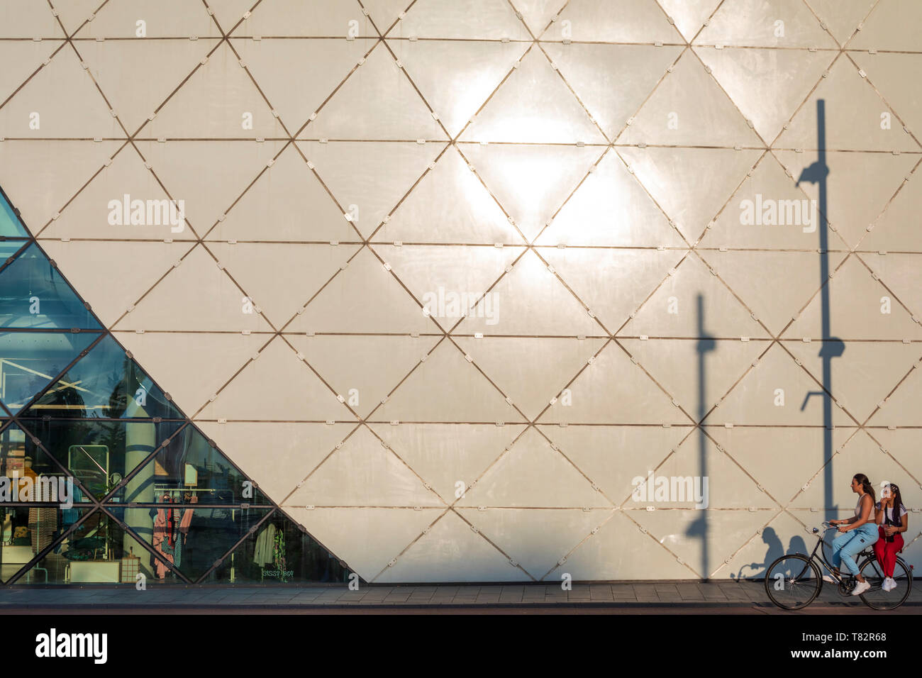 A couple of young women share a bicycle riding past The Blob building in Eindhoven, North Brabant, The Netherlands. Stock Photo