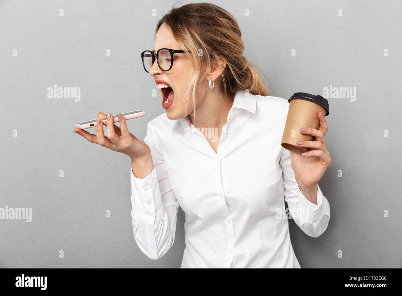 Portrait of angry businesswoman wearing glasses screaming on cell phone and holding coffee in the office isolated over gray background Stock Photo