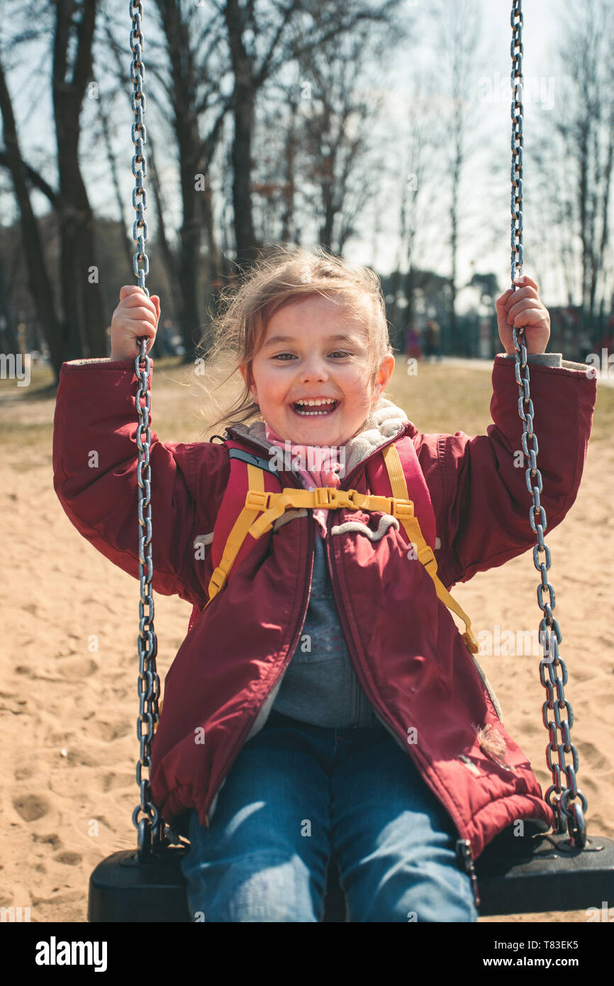 Little smilling happy girl swinging in a park on sunny spring day. Child looking at camera wearing red jacket Stock Photo