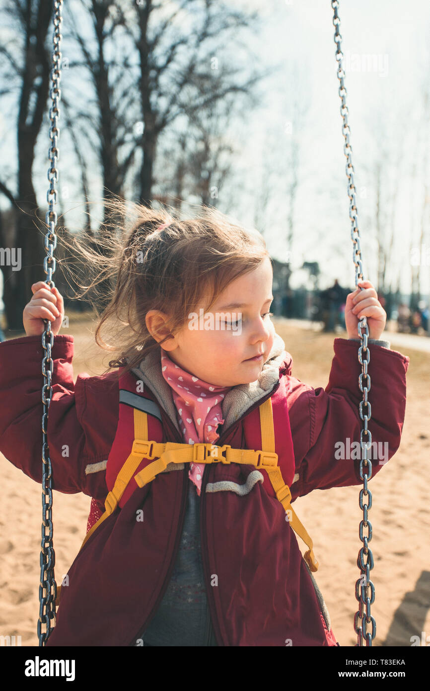 Little cute girl swinging in a park on sunny spring day. Child wearing red jacket Stock Photo