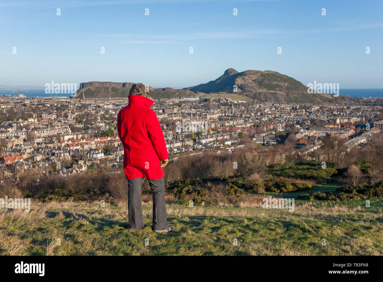 Edinburgh, City of Edinburgh, Scotland. Woman in red coat admiring the view from Blackford Hill over rooftops to Arthur's Seat. Stock Photo