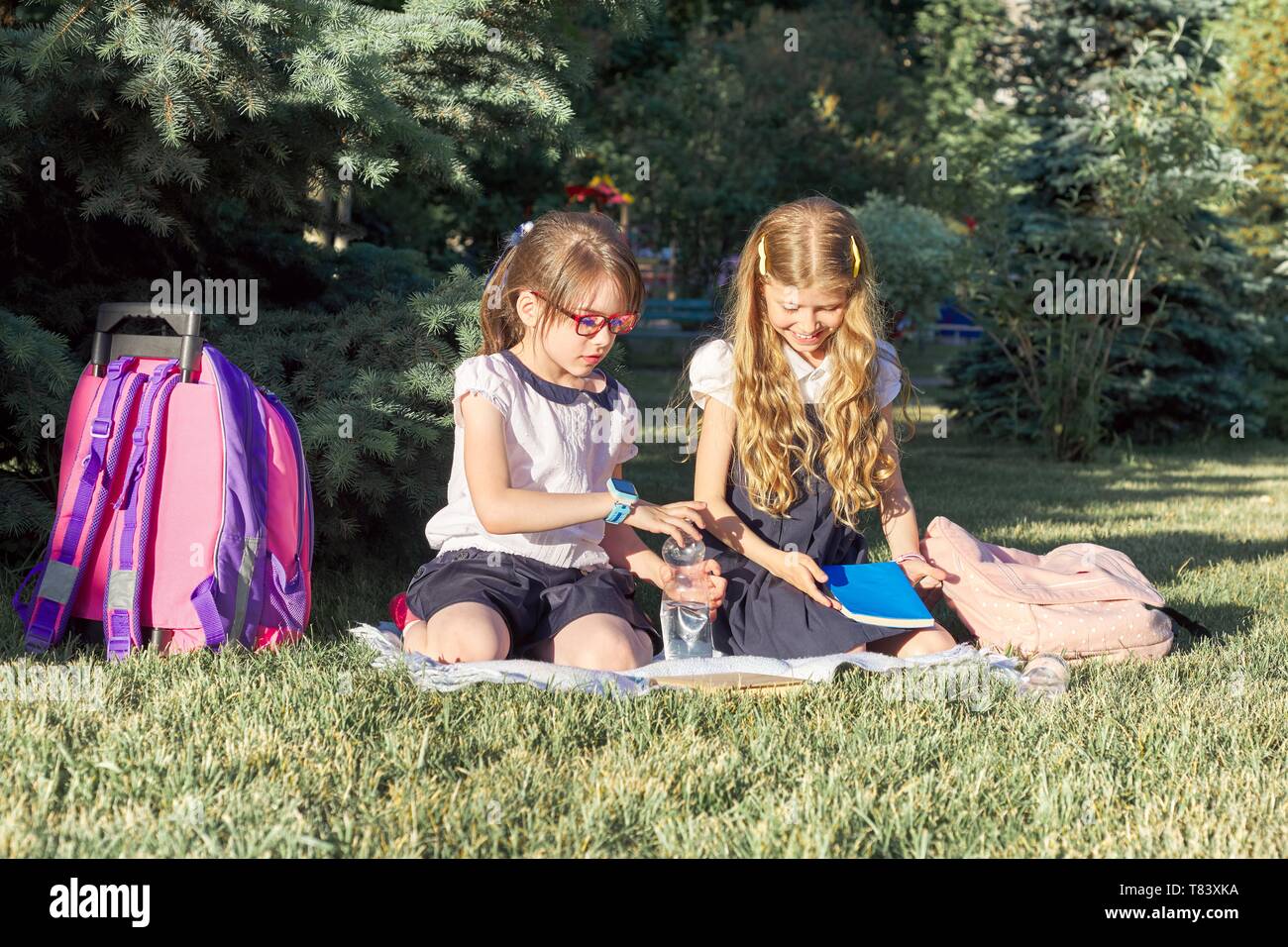 Girls 7, 8 years old in school uniforms with backpacks with books and water bottles sitting in park on grass, hot sunny day Stock Photo