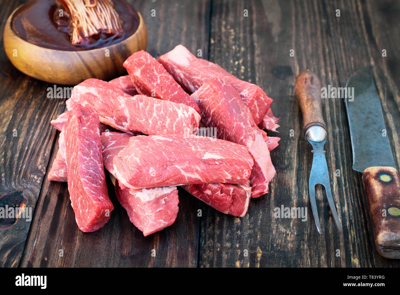 Uncooked raw boneless beef ribs with bowl of bbq sauce and brush in background over top a rustic wood table. Image shot from overhead view. Stock Photo