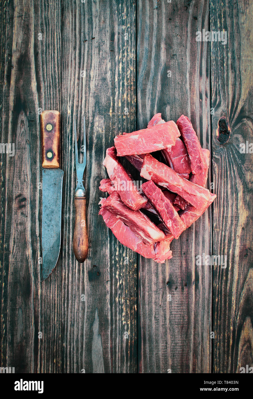 Uncooked raw boneless beef ribs with vintage meat fork and butcher's knife over top a rustic wood table / background. Image shot from overhead view. Stock Photo