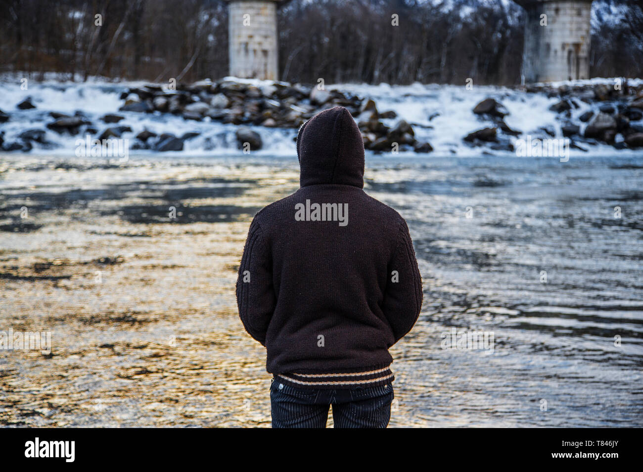 Young man in hood looking out at winter river, rear view, Domodossola, Piemonte, Italy Stock Photo