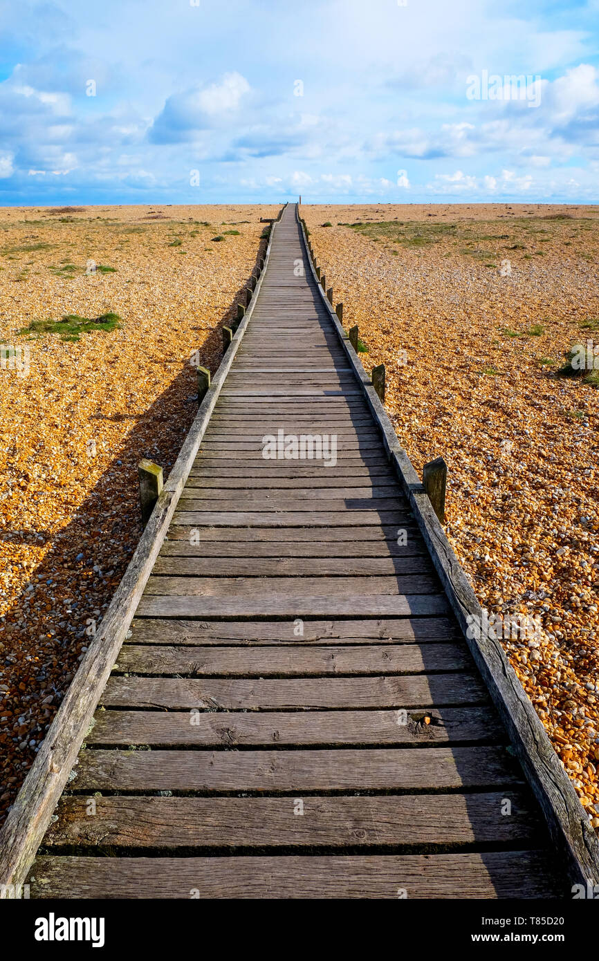 a long straight wooden decking pathway with a diminshing perspective on a pebble beach, the pathway starts large in the foreground centre of the image Stock Photo