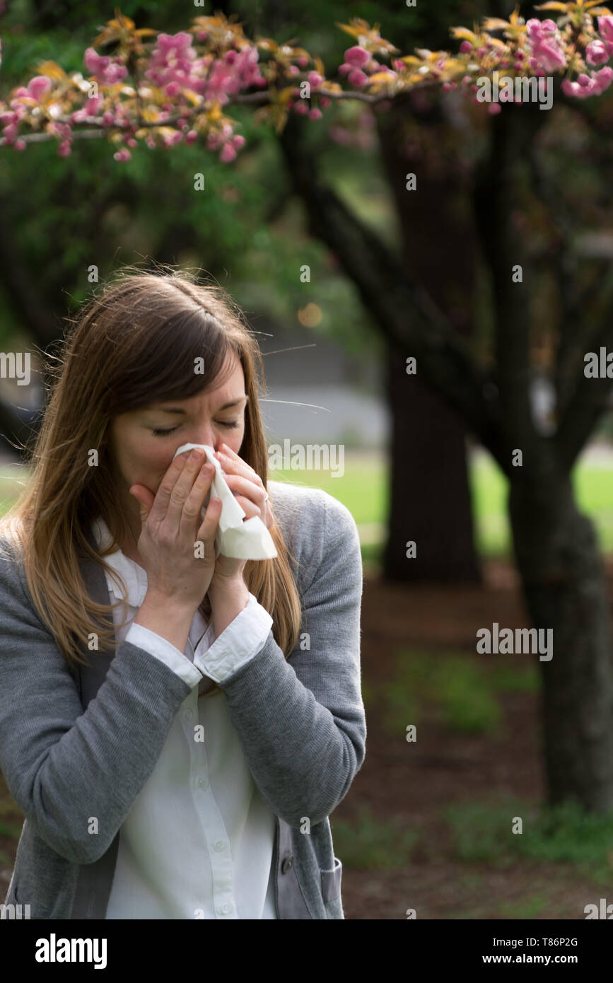 Millennial woman sneezing in the park surrounded by flowers - it looks like allergies! Stock Photo