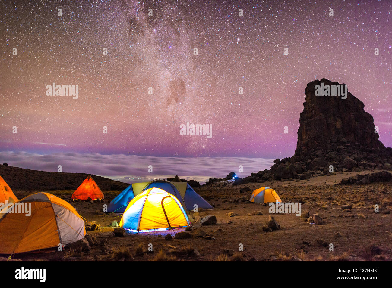 MT KILIMANJARO, Tanzania — The brilliantly clear night sky at Lava Tower Camp (15,215 feet), with Lava Tower at right, clouds in the distance, and the Milky Way. Mt Kilimanjaro's Lemosho Route. Far beyond the tents you can see lights from a town far below. The band of light running near vertically through the sky is the Milky Way. Stock Photo
