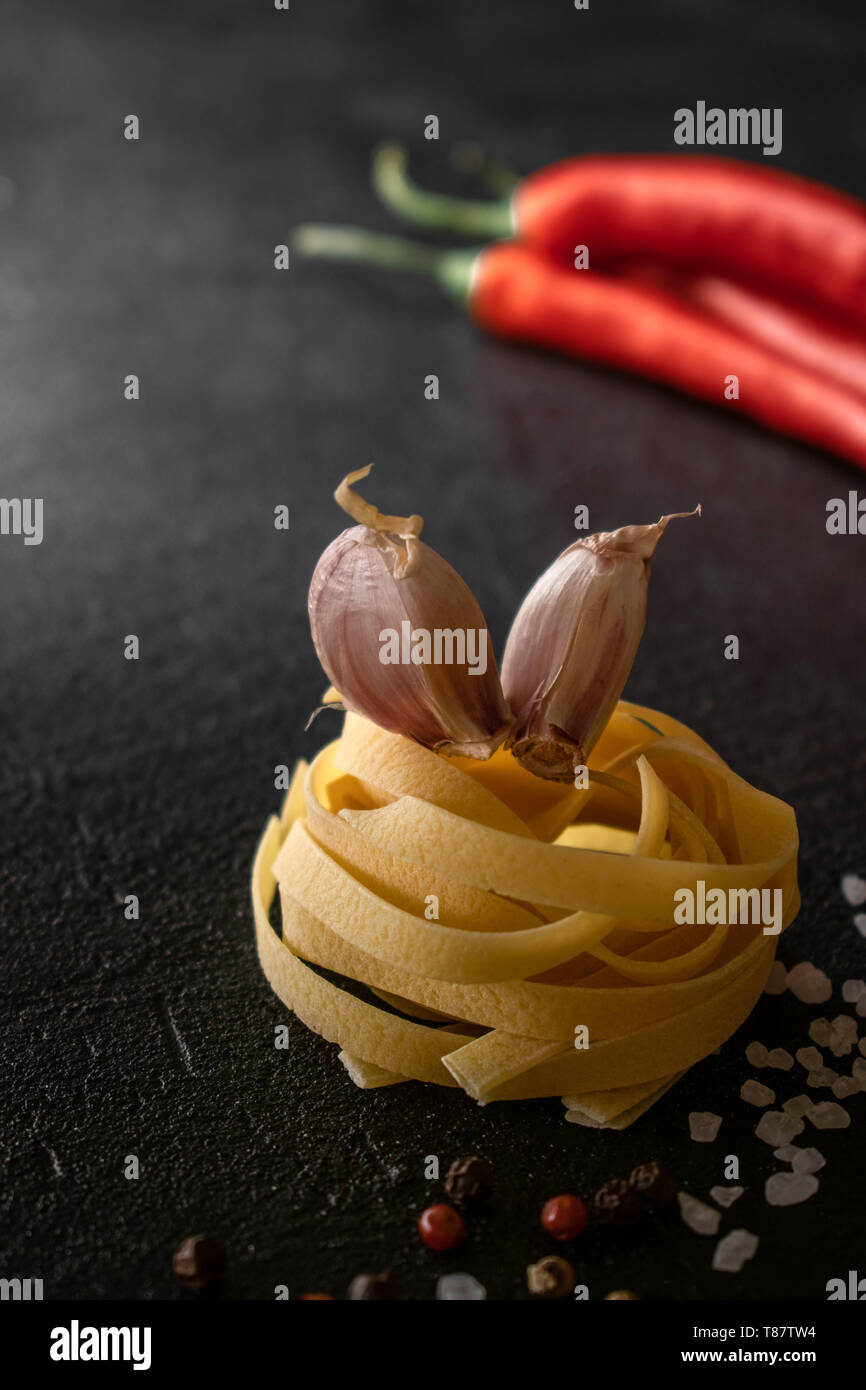 on a black background raw fettuccine paste there are two cloves of garlic, on the side there is sea salt sprinkled, a mixture of peppers, Chilli peppe Stock Photo