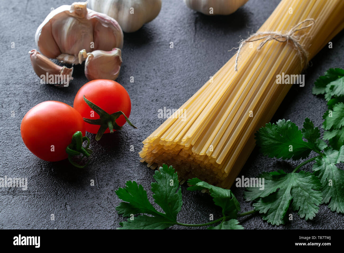 uncooked spaghetti pasta tied up with rope from burlap with cherry tomatoes, garlic heads and fresh branches of green cilantro on black background Stock Photo