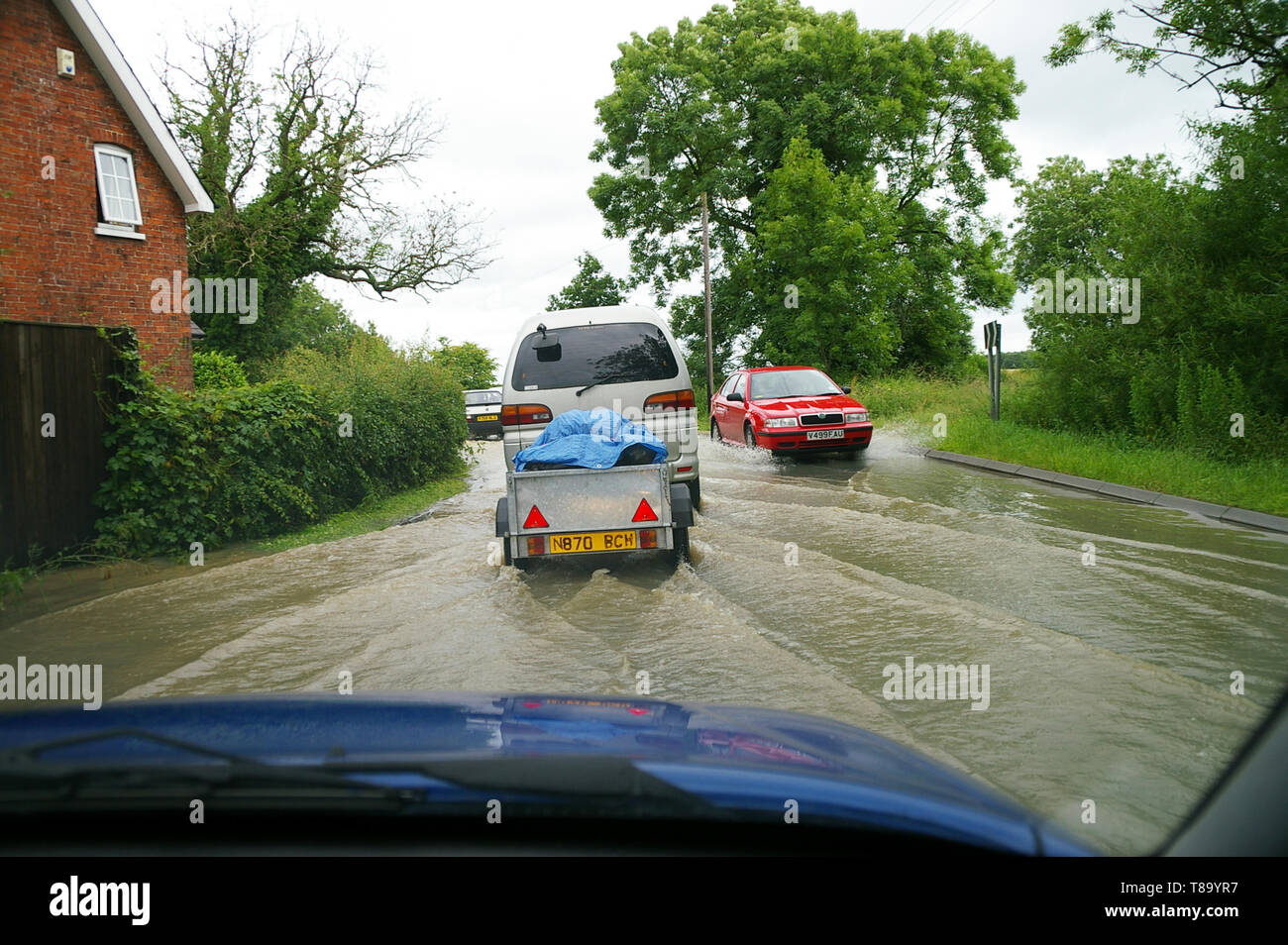 Driving through flooded road with cars. Driver's view from inside car driven through rainwater flood in Lincolnshire country road near property. Stock Photo