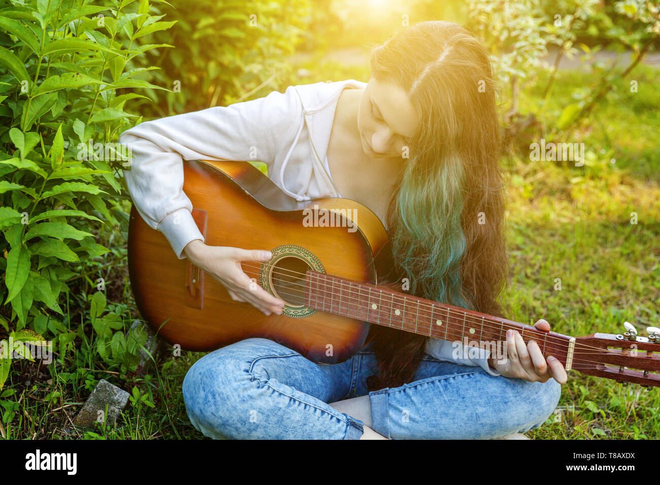 Young hipster woman sitting in grass and playing guitar on park or ...