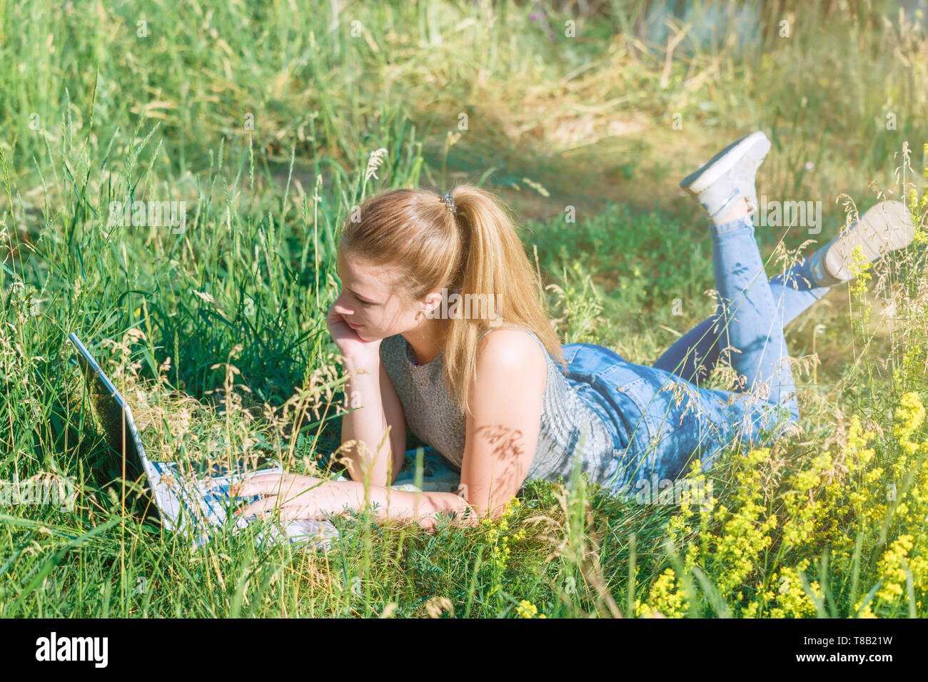 Happy young girl lying on the grass and looking at the laptop monitor ...