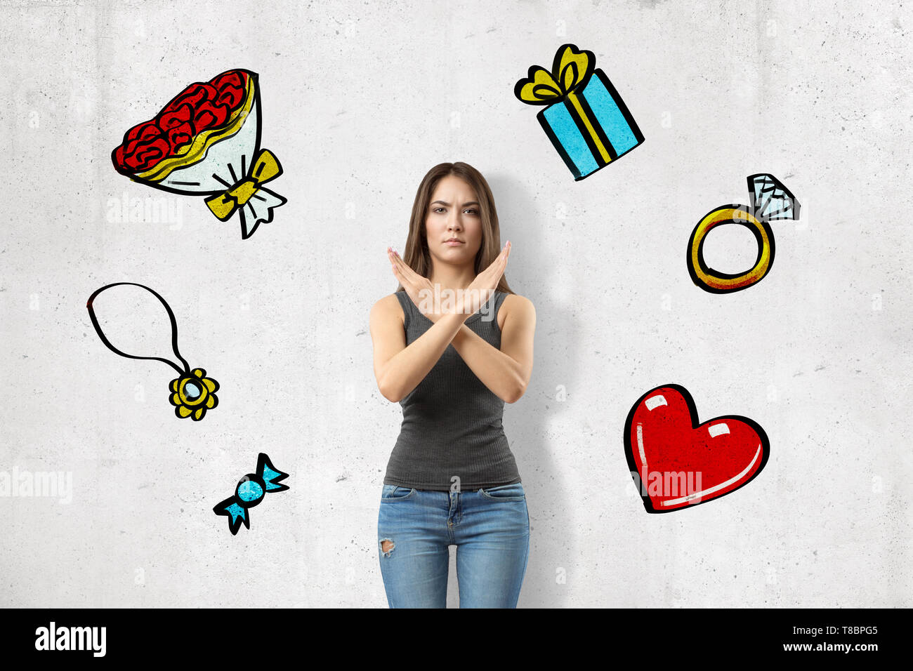 Front view of young angry girl, crossing arms in stop gesture, standing at wall with images of ring, gift, bouquet, heart, candy and jewelry on it. Stock Photo