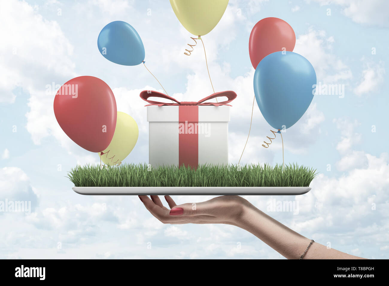 Side view of woman's hand holding digital tablet with green grass on screen, gift box standing on top and colorful balloons flying around. Stock Photo
