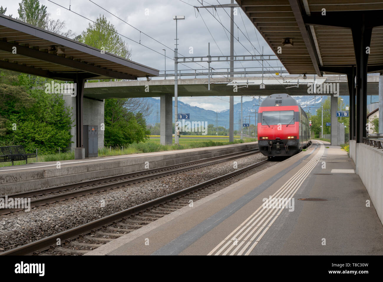red train speeds through a small rural train station with nobody on the platform in the Swiss countryside Stock Photo