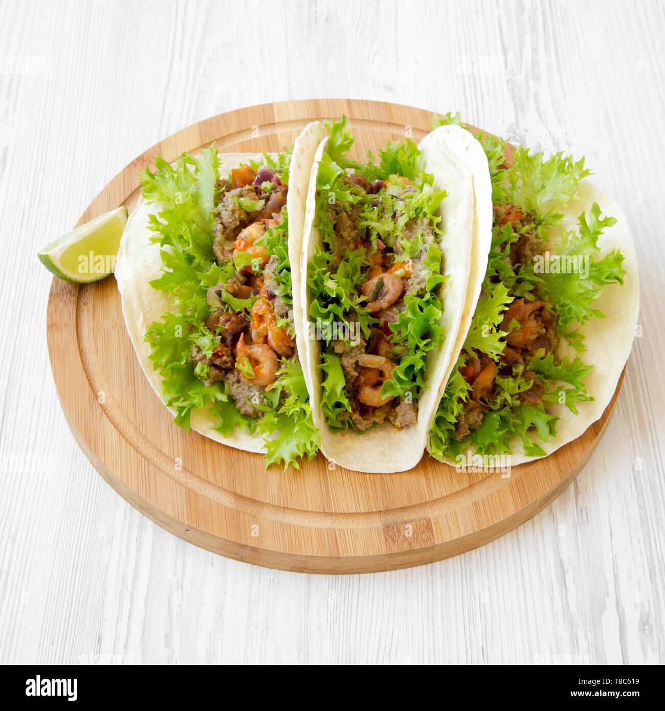 Shrimp tacos on a round bamboo board over white wooden background, side view. Mexican cuisine. Close-up. Stock Photo