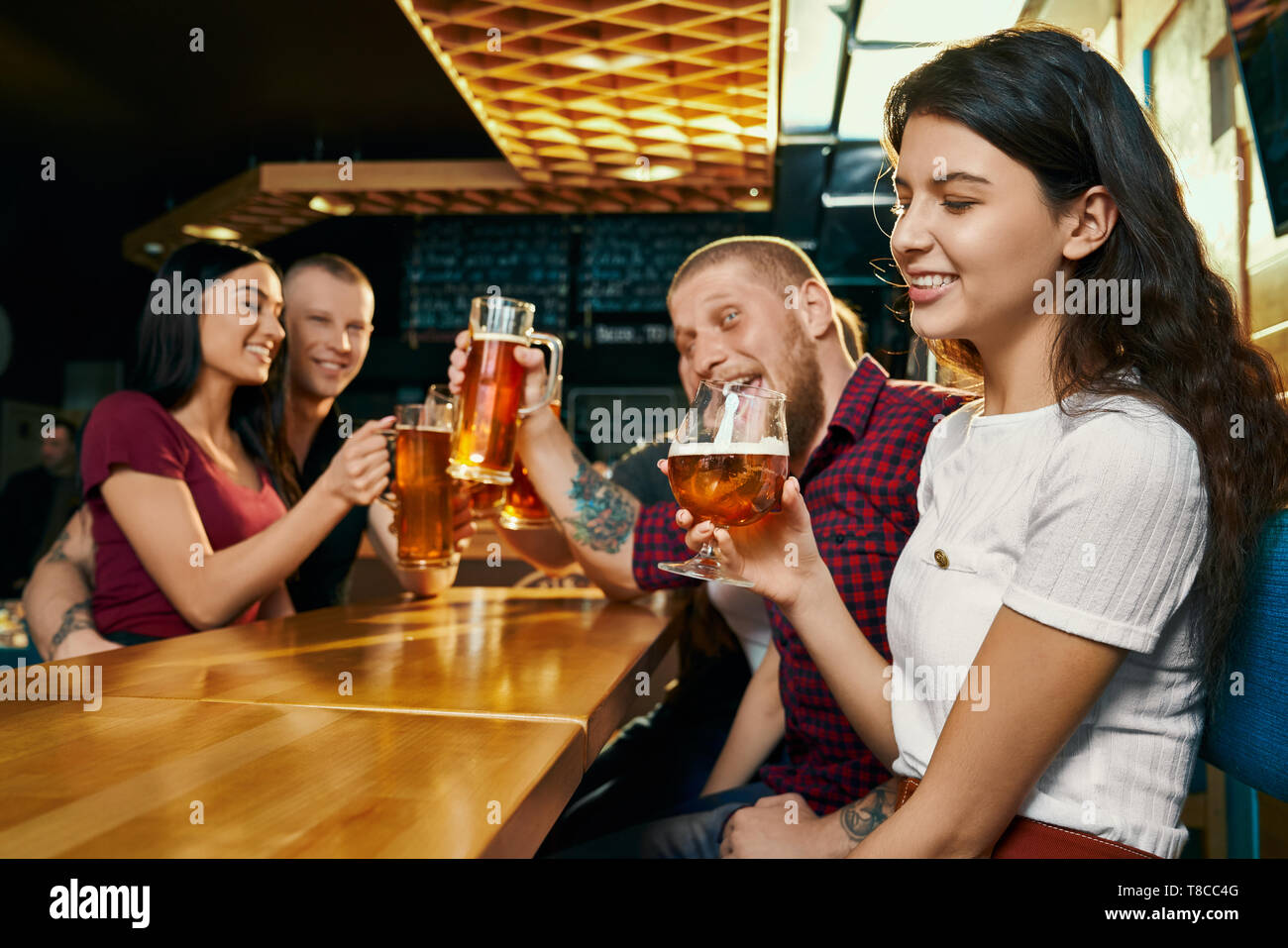 Side view of young smiling brunette enjoying free time with happy friends and drinking beer in bar. Cheerful company drinking alcohol, talking and laughing in pub. Concept of beverage and fun. Stock Photo