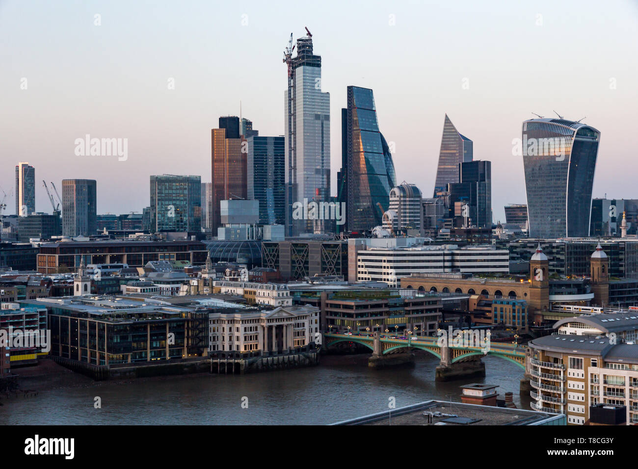 The bold and imposing City of London skyscrapers reflect a golden colour at sunset with a clear sky and the River Thames in the foreground Stock Photo