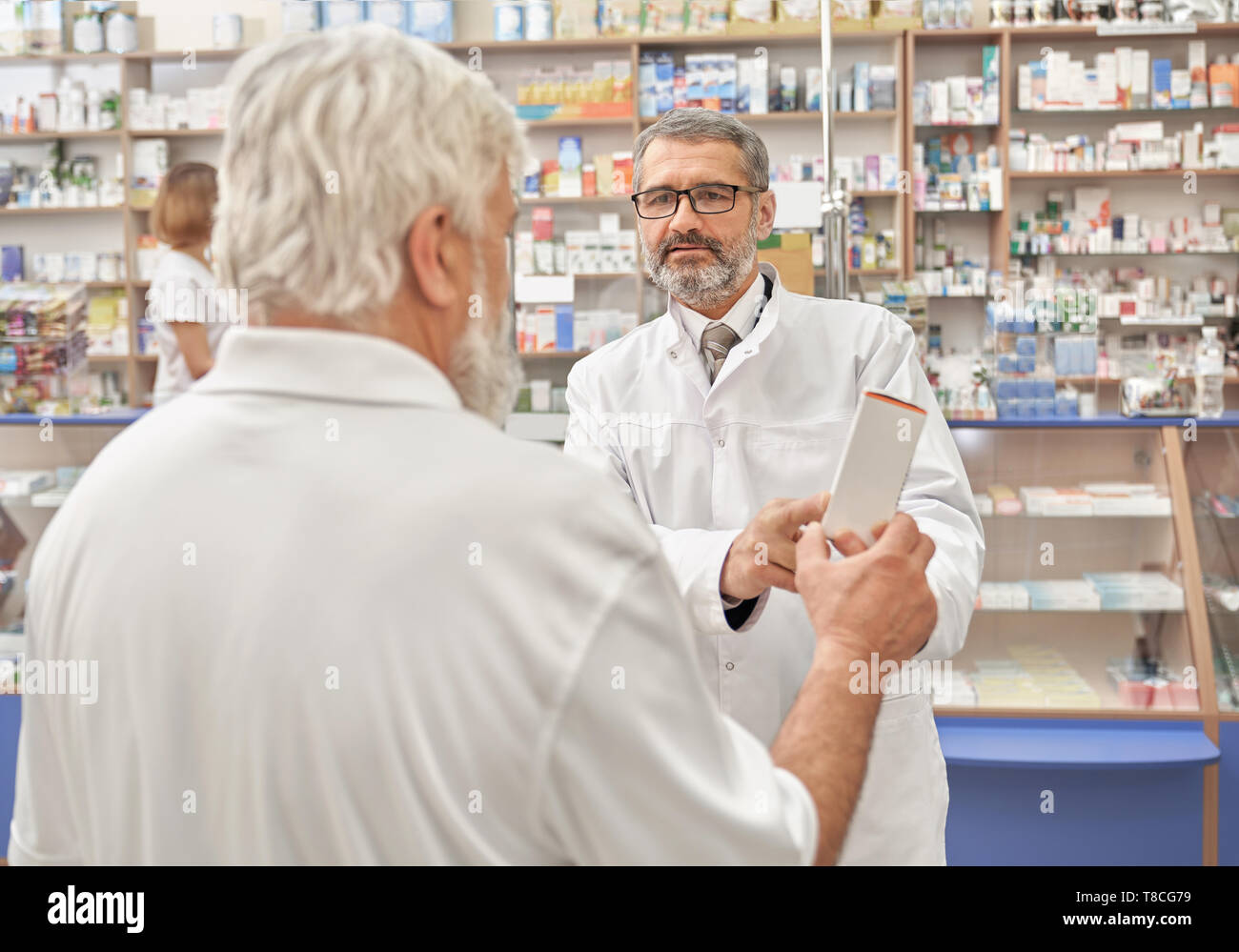 Pensioner talking with doctor about medicines and treatment of disease. Specialist explaining, helping elderly man with choice of medicament. Doctor wearing in white lab coat. Stock Photo