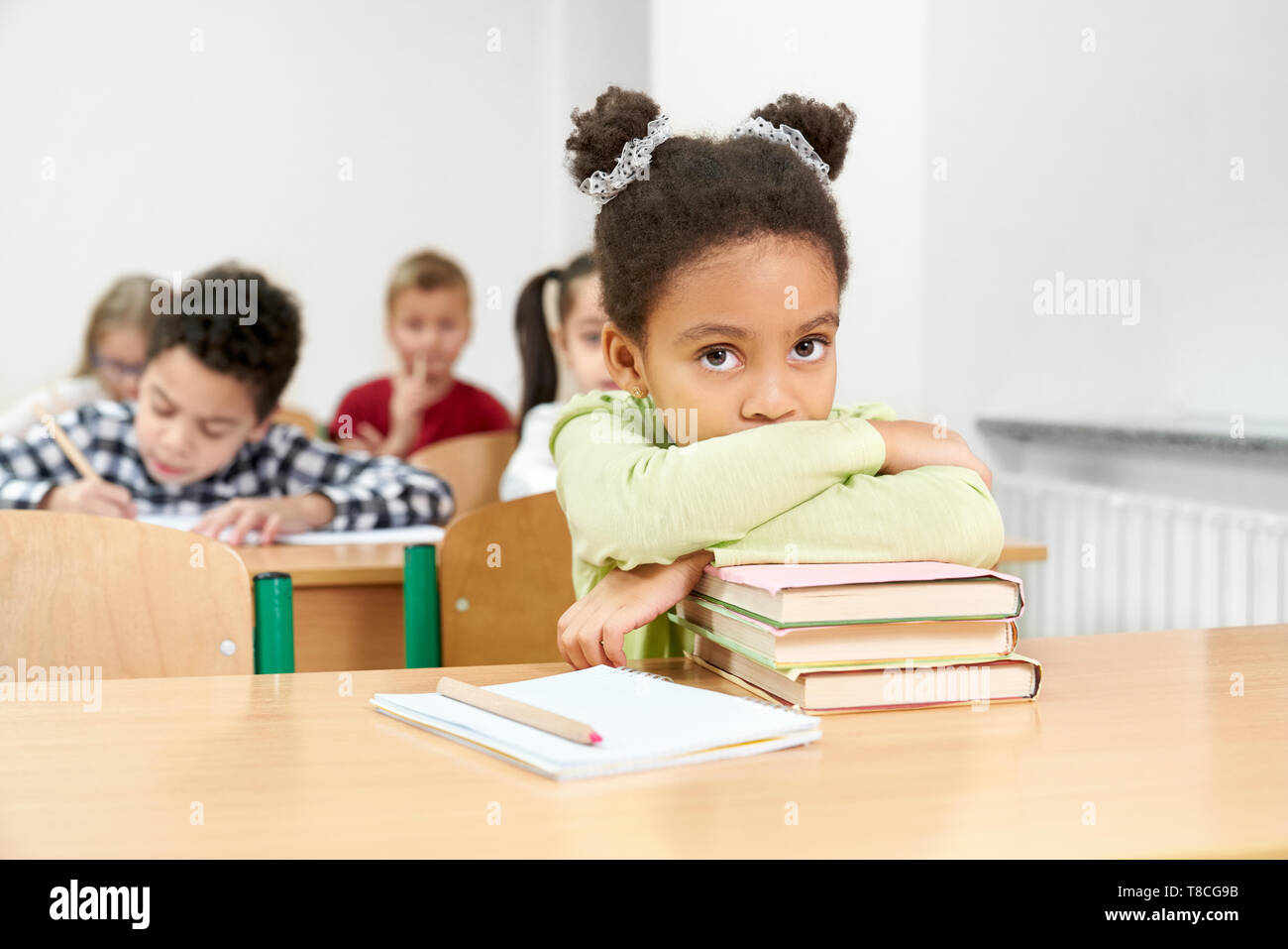 Pretty schoolgirl sitting at desk with copybook and pen in classroom, leaning on books. Cute child posing, looking at camera. Group of pupils sitting at tables in class, writing.  Stock Photo