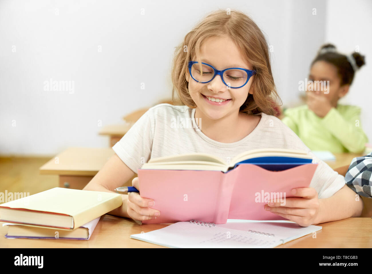 Pretty, happy schoolgirl in blue glasses sitting at desk, holding book, reading, smiling. Beautiful, cheerful teenager studying in school looking in book, posing in classroom. Stock Photo