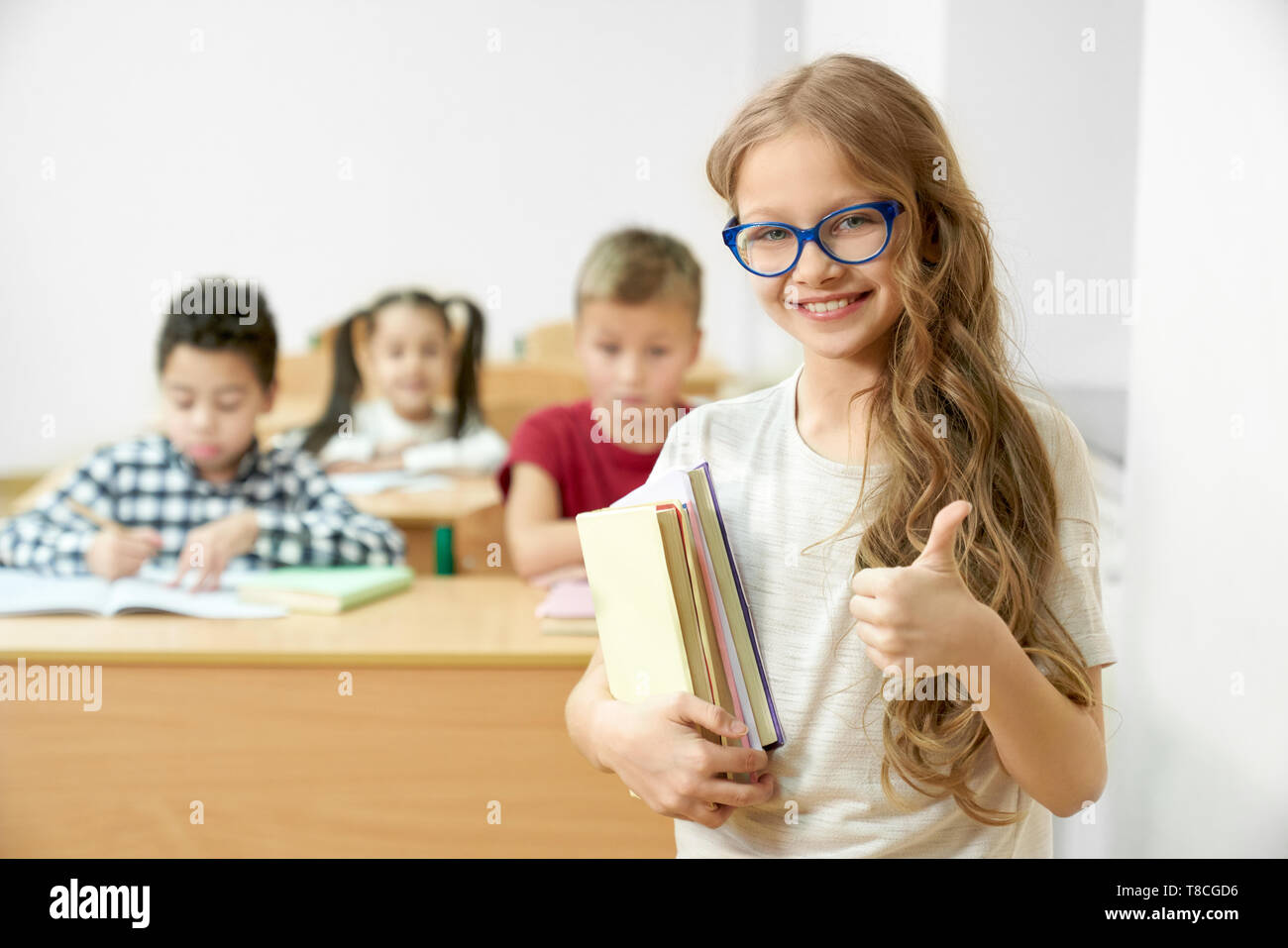 Cheerful, positive, cute schoolgirl standing in classroom, holding books and showing thumb up. Pretty child of primary school in glasses looking at camera, posing. Stock Photo