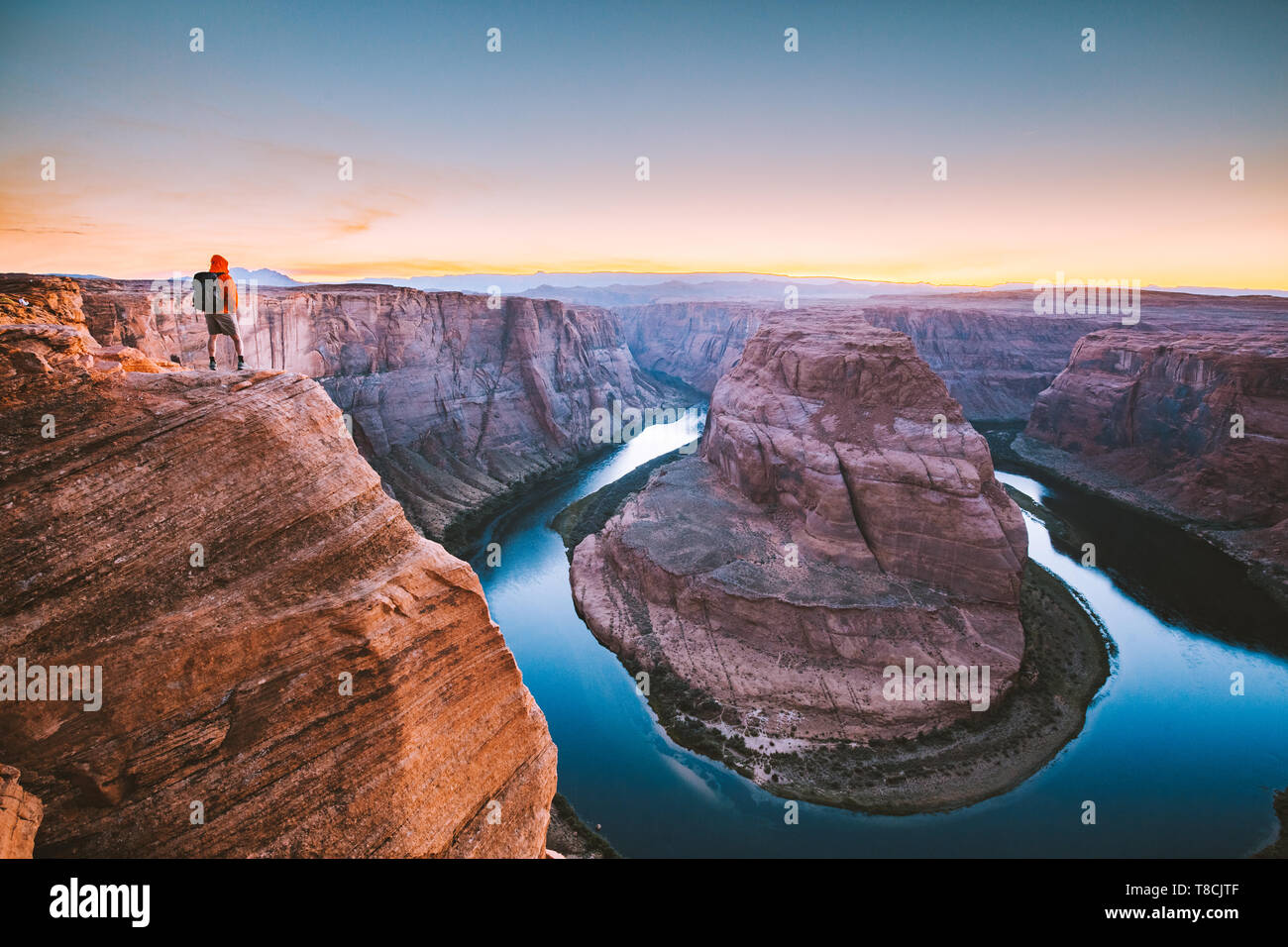 A male hiker is standing on steep cliffs enjoying the beautiful view of Colorado river flowing at famous Horseshoe Bend at sunset, Arizona, USA Stock Photo